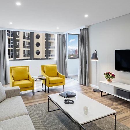 Meriton Suites Bondi Junction Сидней Экстерьер фото The photo shows a modern, well-lit living room. It features a light gray sofa with a textured fabric. There are two bright yellow armchairs positioned near a window, which allows natural light to enter the space. The room has light gray curtains and 