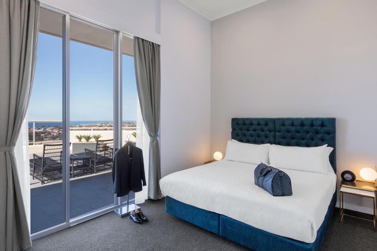 Meriton Suites Bondi Junction Сидней Экстерьер фото The photo shows a stylish bedroom featuring a large bed with a tufted blue headboard. There's a neatly organized outfit consisting of a suit hanging on a stand, along with shoes placed on the floor. The room has large windows that provide a view of t