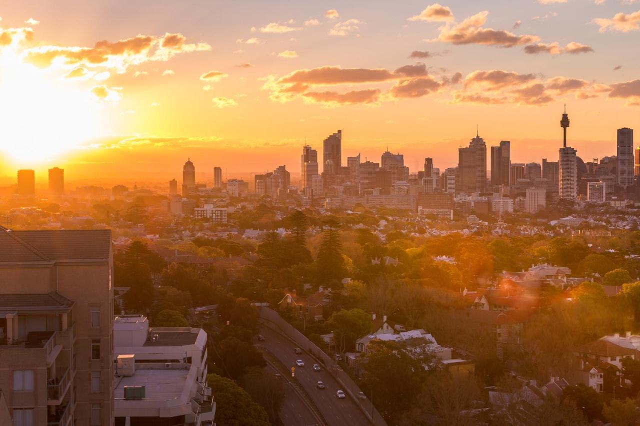 Meriton Suites Bondi Junction Сидней Экстерьер фото The photo shows a panoramic view of a city skyline at sunset. The sun is low on the horizon, casting a warm golden light across the buildings. Tall skyscrapers are silhouetted against the colorful sky, which features hues of orange, pink, and yellow,