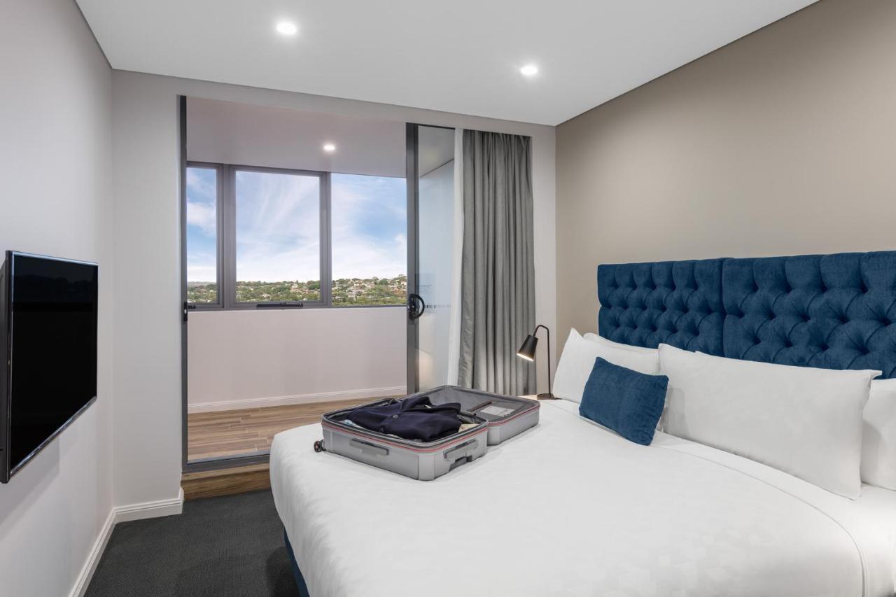 Meriton Suites Bondi Junction Сидней Экстерьер фото The photo shows a modern hotel room featuring a double bed with a tufted blue headboard. There is a neatly arranged suitcase placed open on the bed, indicating that someone may be unpacking. The room has light-colored walls and a window allowing natu