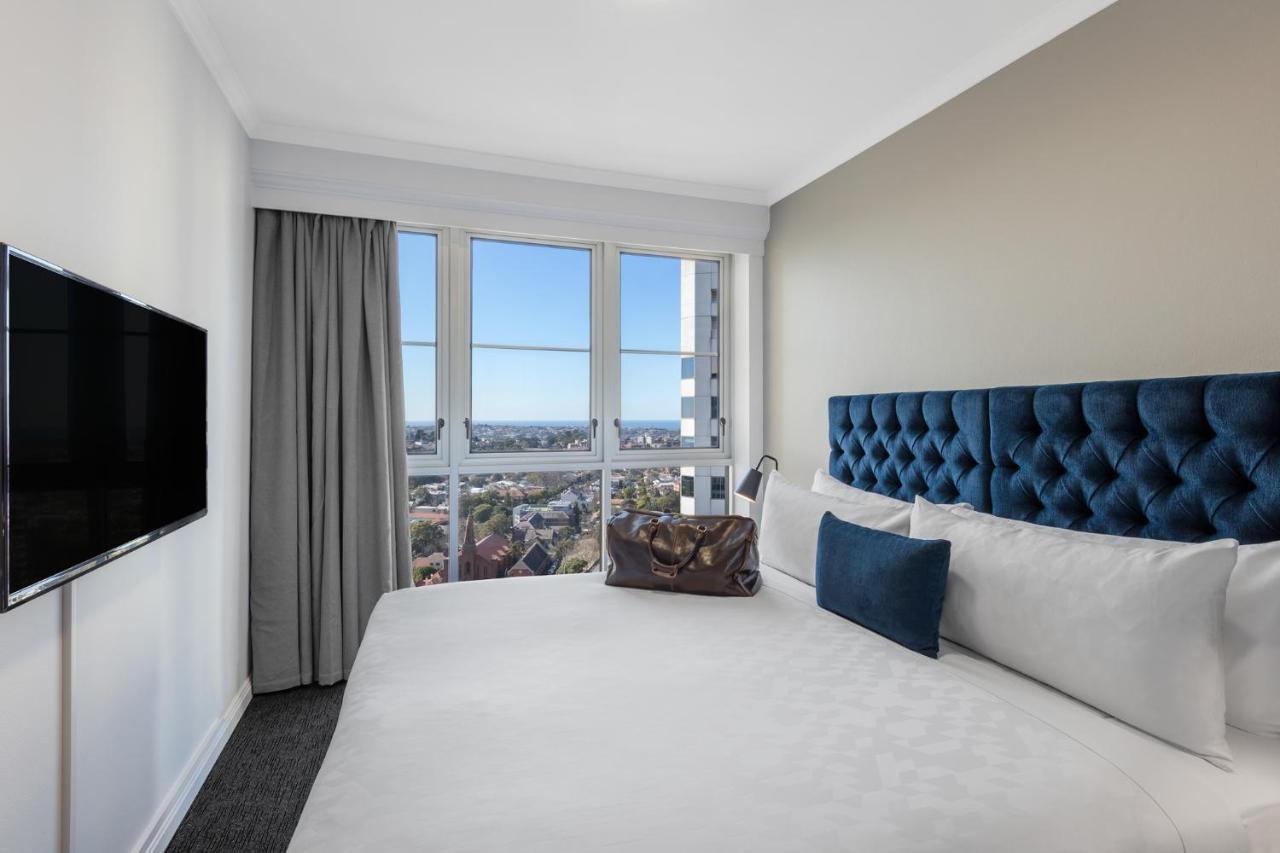 Meriton Suites Bondi Junction Сидней Экстерьер фото The photo shows a modern hotel room featuring a large bed with a tufted blue headboard. Beside the bed, there is a stylish bag placed on the bed. The room has large windows that provide a view of the outside, allowing natural light to fill the space.