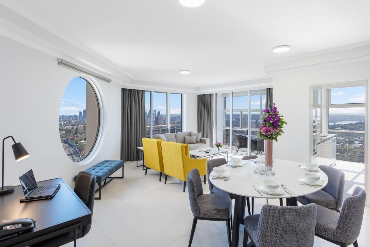 Meriton Suites Bondi Junction Сидней Экстерьер фото The photo shows a modern and bright interior space, likely a living room or dining area. It features large windows that offer a panoramic view of the city skyline. The furnishings include a round dining table set for several people, with plates and g