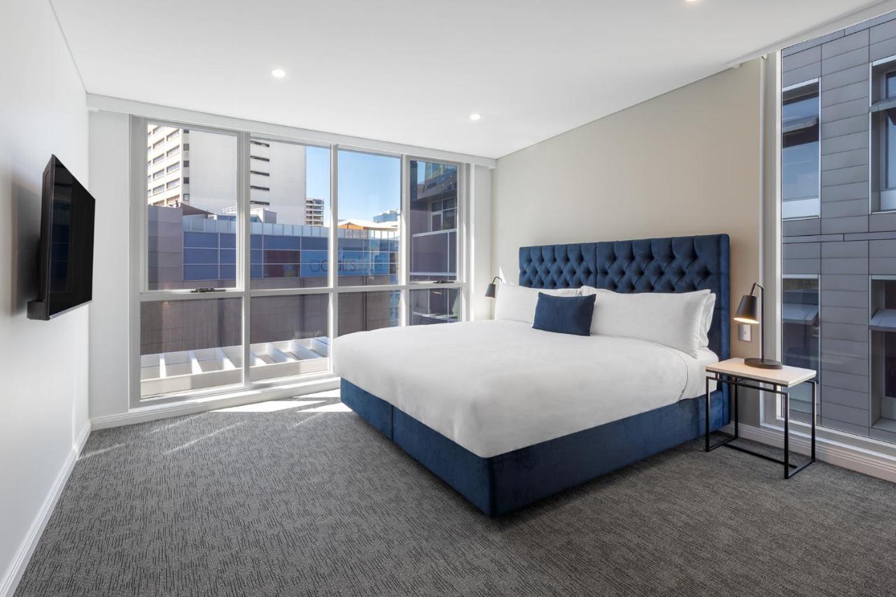 Meriton Suites Bondi Junction Сидней Экстерьер фото The image shows a modern hotel room with a large, comfortable bed featuring a blue upholstered headboard. The room has large windows that allow natural light to enter, providing a good view of the surrounding buildings. There is a small bedside table