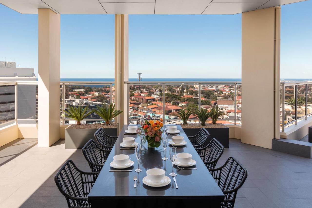 Meriton Suites Bondi Junction Сидней Экстерьер фото The photo shows a modern outdoor dining area situated on a balcony or terrace with a stunning view. There is a long rectangular table set with plates, glasses, and cutlery, ready for a meal. The table is surrounded by sleek, black wicker chairs. In t