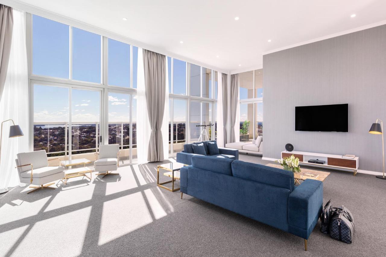 Meriton Suites Bondi Junction Сидней Экстерьер фото The photo shows a bright and modern living room with large floor-to-ceiling windows that provide a view of the outdoors. The space features a blue sofa in the center, complemented by light-colored armchairs nearby. There are light curtains hanging by