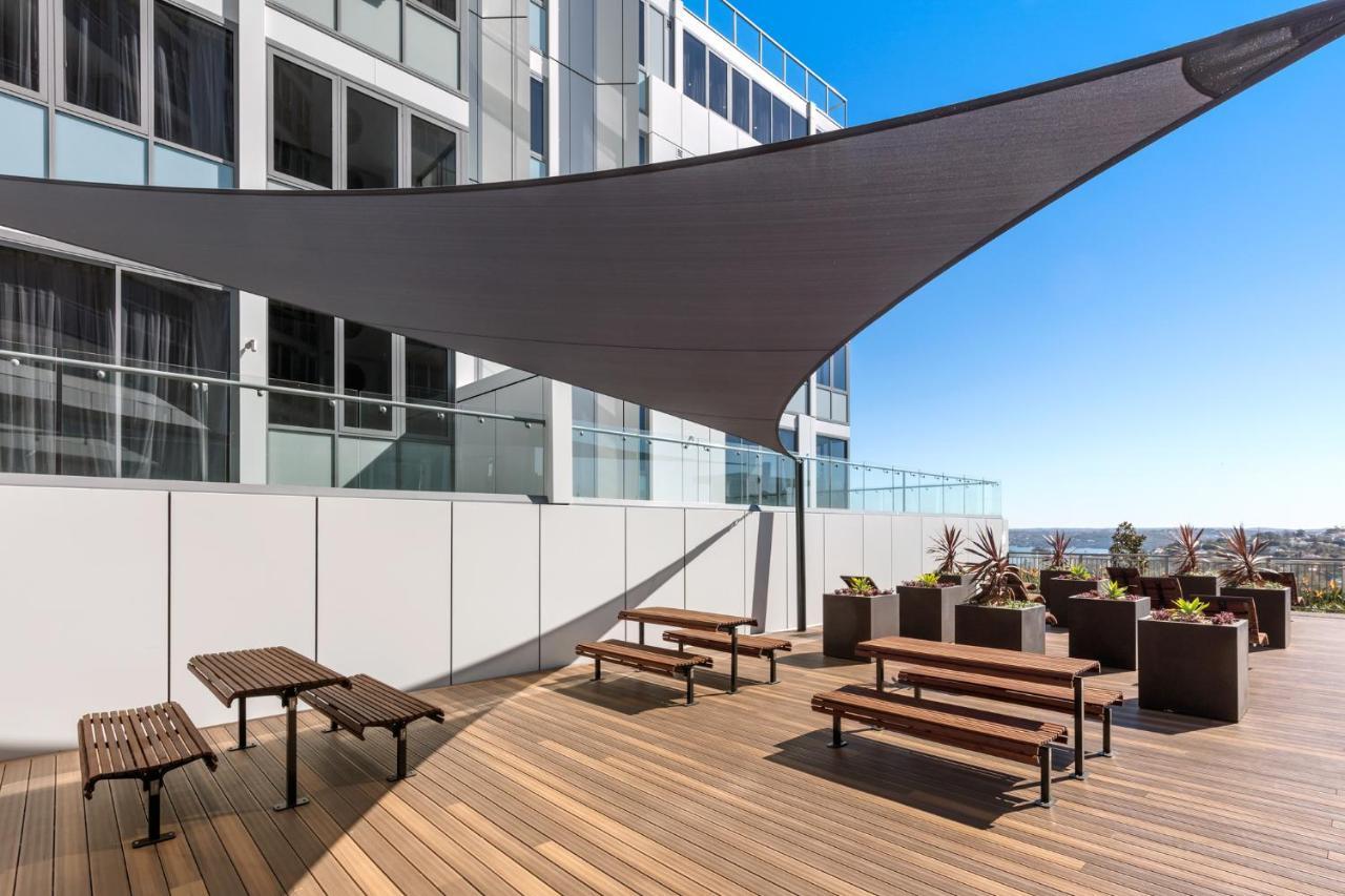 Meriton Suites Bondi Junction Сидней Экстерьер фото The photo shows a modern outdoor terrace area on a building. It features several wooden benches arranged on a wooden deck. There are large planters with greenery, adding some decorative elements. A large triangular gray shade sail is positioned above