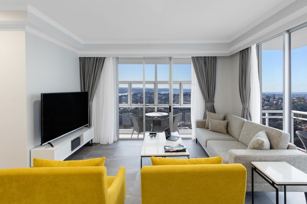 Meriton Suites Bondi Junction Сидней Экстерьер фото The photo depicts a modern living room space with large windows that offer a view of the outside. The decor is minimalist and stylish, featuring a light color palette. 

In the center, there are two bright yellow chairs facing each other. A sleek, re