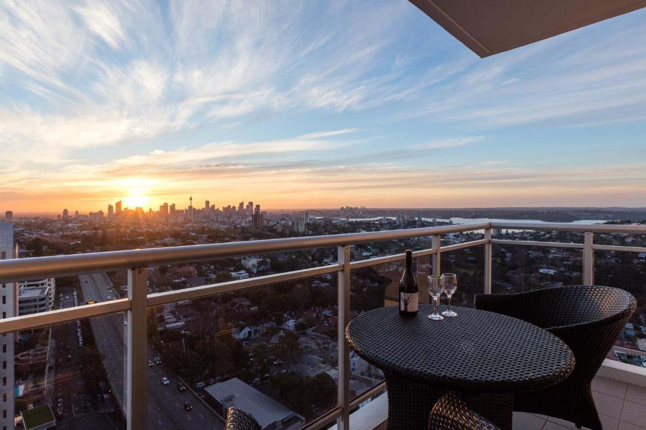 Meriton Suites Bondi Junction Сидней Экстерьер фото The image shows a scenic view from a balcony or terrace overlooking a cityscape during sunset. In the foreground, there is a small round table with two glasses and a bottle of wine, suggesting a romantic or relaxing setting. The sky is filled with vi