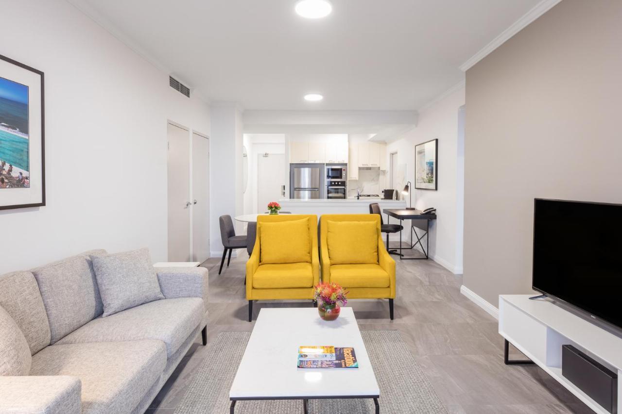 Meriton Suites Bondi Junction Сидней Экстерьер фото The photo shows a modern living space with a light and airy design. In the foreground, there are two bright yellow armchairs flanking a white coffee table, which has a small vase of flowers and a magazine on it. To the left, there is a light gray sof