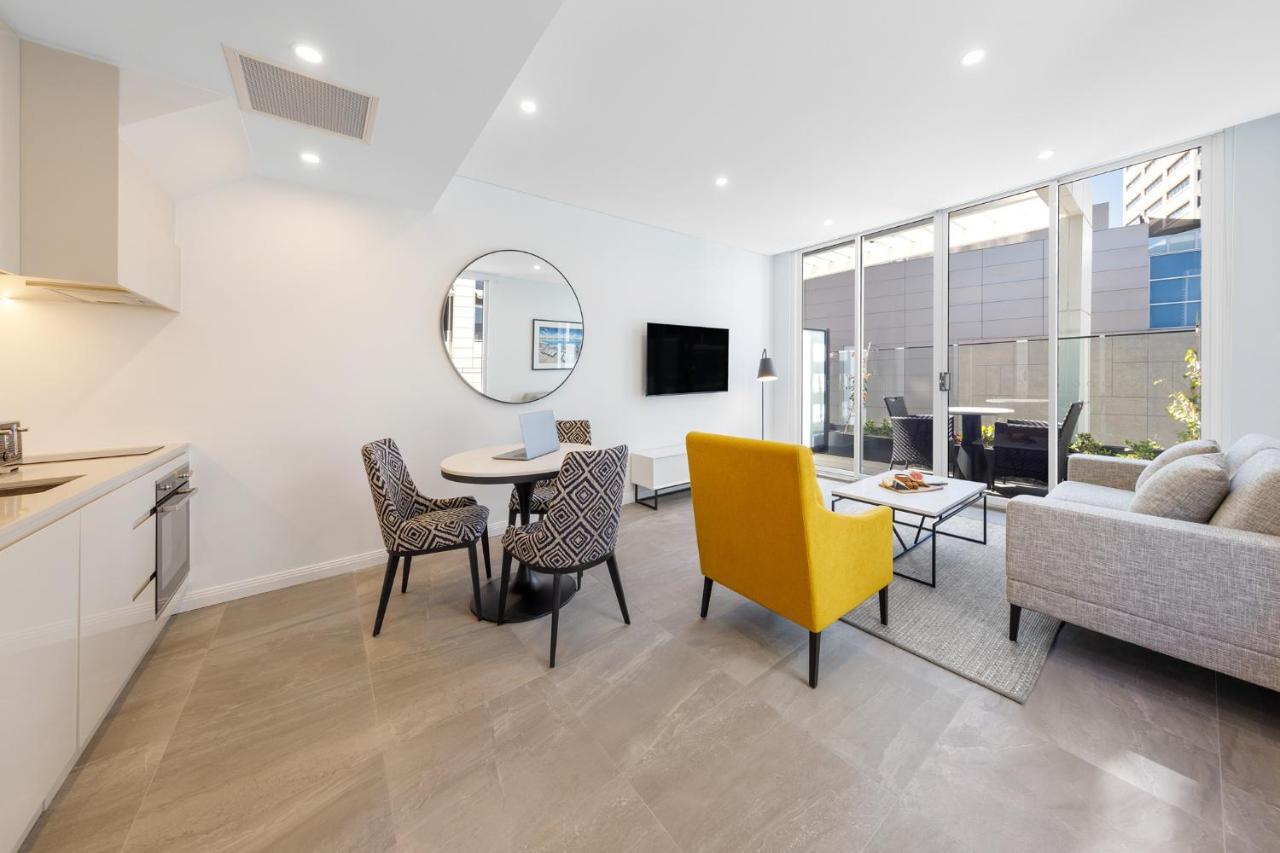 Meriton Suites Bondi Junction Сидней Экстерьер фото The photo shows a modern and stylish living space. On one side, there is a kitchen area with sleek cabinetry, and next to it, a round dining table surrounded by three patterned chairs. In the center of the room, there is a gray sofa accompanied by a 