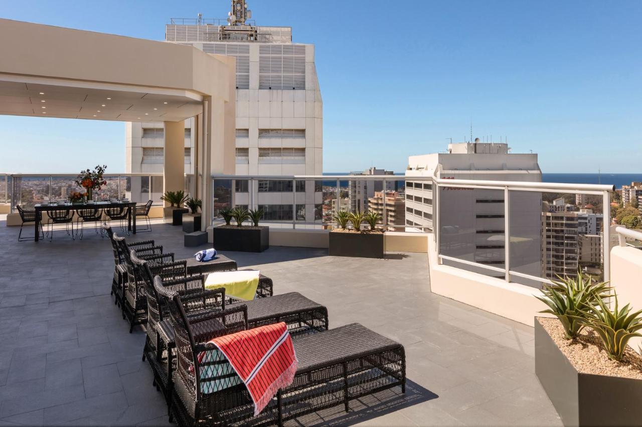 Meriton Suites Bondi Junction Сидней Экстерьер фото The photo shows a rooftop terrace with a modern design. There are several outdoor seating arrangements featuring black wicker lounge chairs. Some of the chairs have colorful blankets draped over them. The terrace is surrounded by a railing and has po