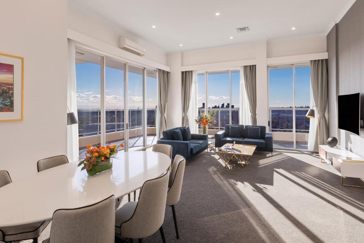 Meriton Suites Bondi Junction Сидней Экстерьер фото The photo shows a modern and stylish living space with large windows allowing natural light to fill the room. In the foreground, there is a round dining table with several chairs around it, adorned with a floral centerpiece. The seating area features