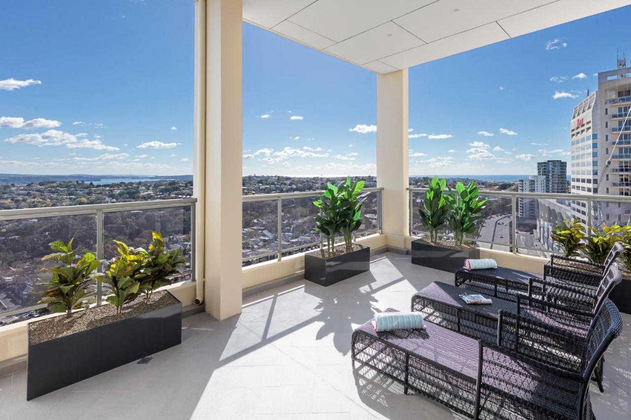 Meriton Suites Bondi Junction Сидней Экстерьер фото The photo shows a modern balcony or terrace area with a stunning view. There are several planters with green plants placed along the edge. The seating area features lounge chairs in a contemporary design, likely made of woven materials. The space app