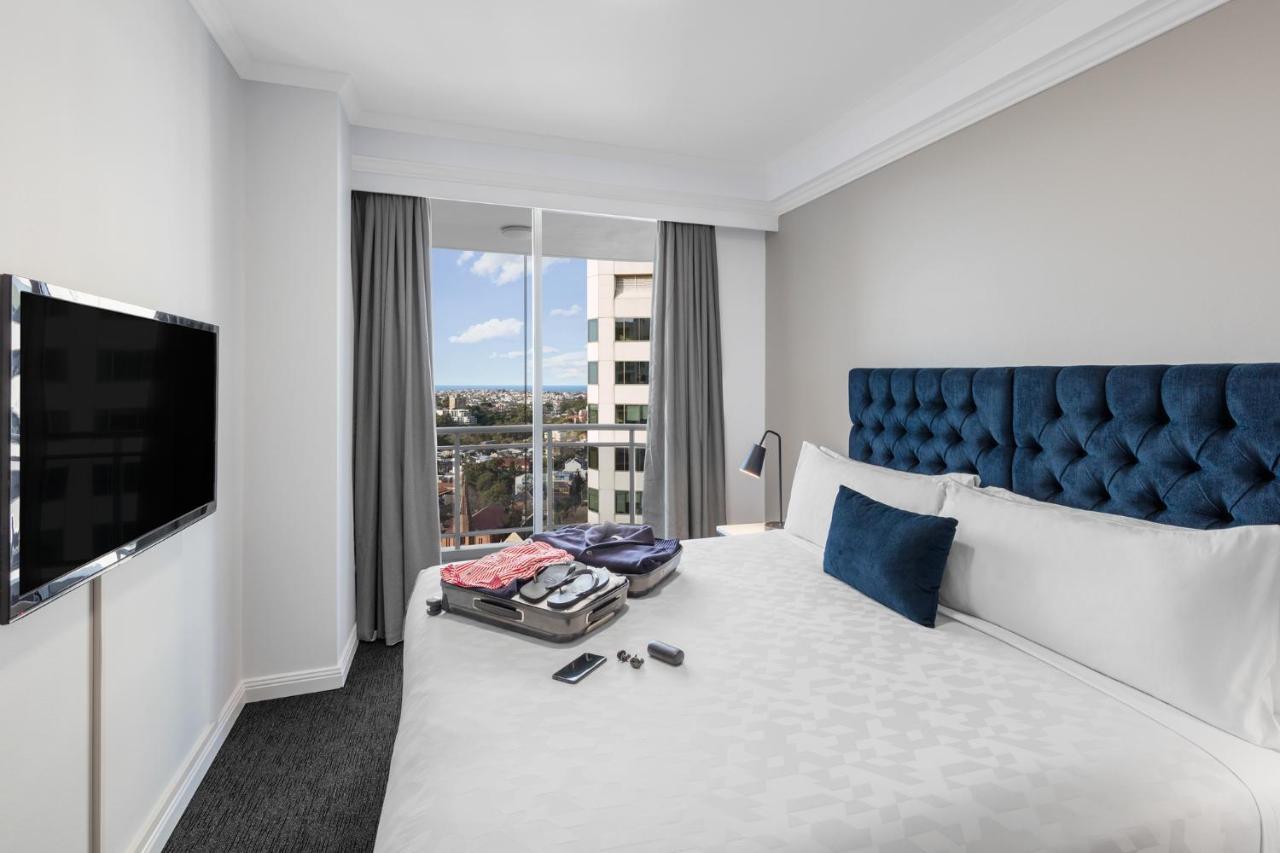 Meriton Suites Bondi Junction Сидней Экстерьер фото The photo shows a modern hotel room featuring a neatly made bed with a blue tufted headboard and a white duvet. There's a small decorative pillow on the bed, and a piece of luggage is placed at the foot with clothes partially unpacked. A television i