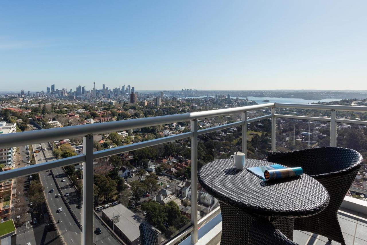 Meriton Suites Bondi Junction Сидней Экстерьер фото The photo shows a balcony with a view overlooking a cityscape and a body of water in the distance. There is a round table with a woven design and two chairs, one of which has a mug on it. On the table, there’s also a rolled-up magazine or document. T