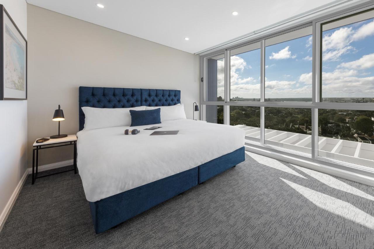 Meriton Suites Bondi Junction Сидней Экстерьер фото The photo shows a modern and stylish hotel room. It features a comfortable queen-size bed with a blue tufted headboard. The bed is neatly made with white linens and pillows. Next to the bed, there is a small bedside table with a desk lamp. 

On the f
