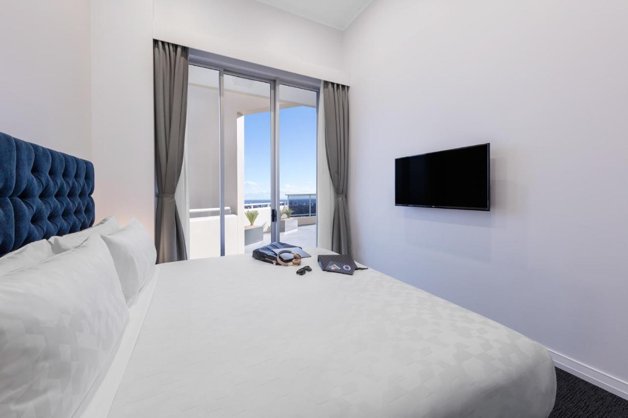 Meriton Suites Bondi Junction Сидней Экстерьер фото The photo shows a modern hotel room with a large, comfortable bed featuring a blue tufted headboard. There are light-colored walls, and the room is decorated simply. A flat-screen TV is mounted on the wall opposite the bed. The room has large glass d