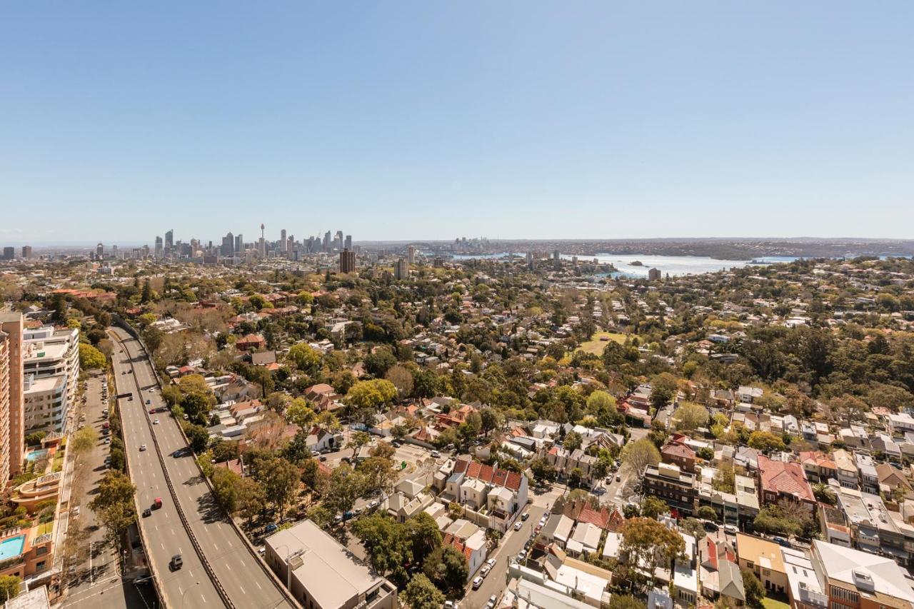 Meriton Suites Bondi Junction Сидней Экстерьер фото The photo captures a panoramic view of a vibrant urban landscape. In the foreground, there are residential areas with various houses and greenery, showcasing a suburban environment. In the background, the skyline of a city can be seen, featuring tall