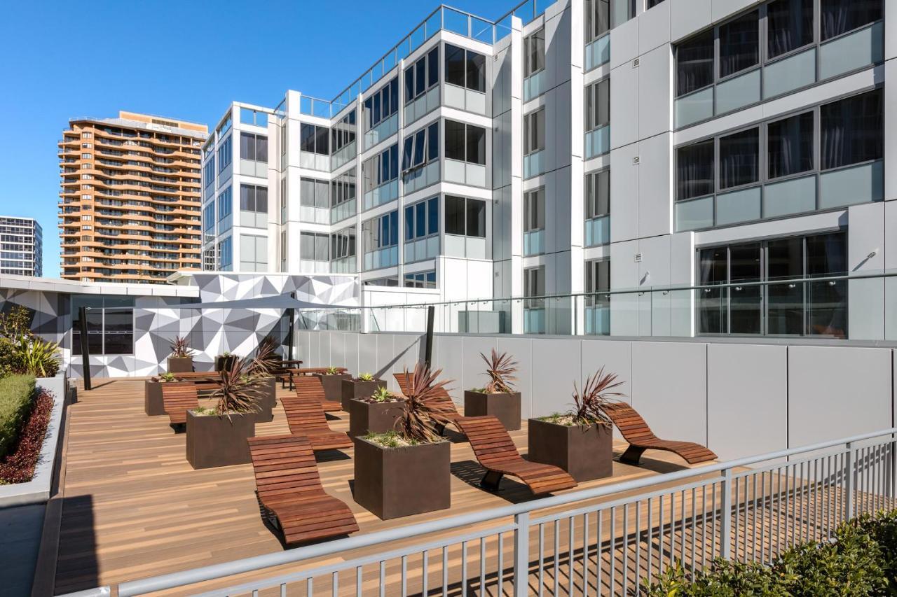 Meriton Suites Bondi Junction Сидней Экстерьер фото The photo shows a modern outdoor space featuring a wooden deck. There are several lounge chairs arranged around large planter boxes, which contain plants. In the background, there are contemporary residential buildings with large windows, and a geome
