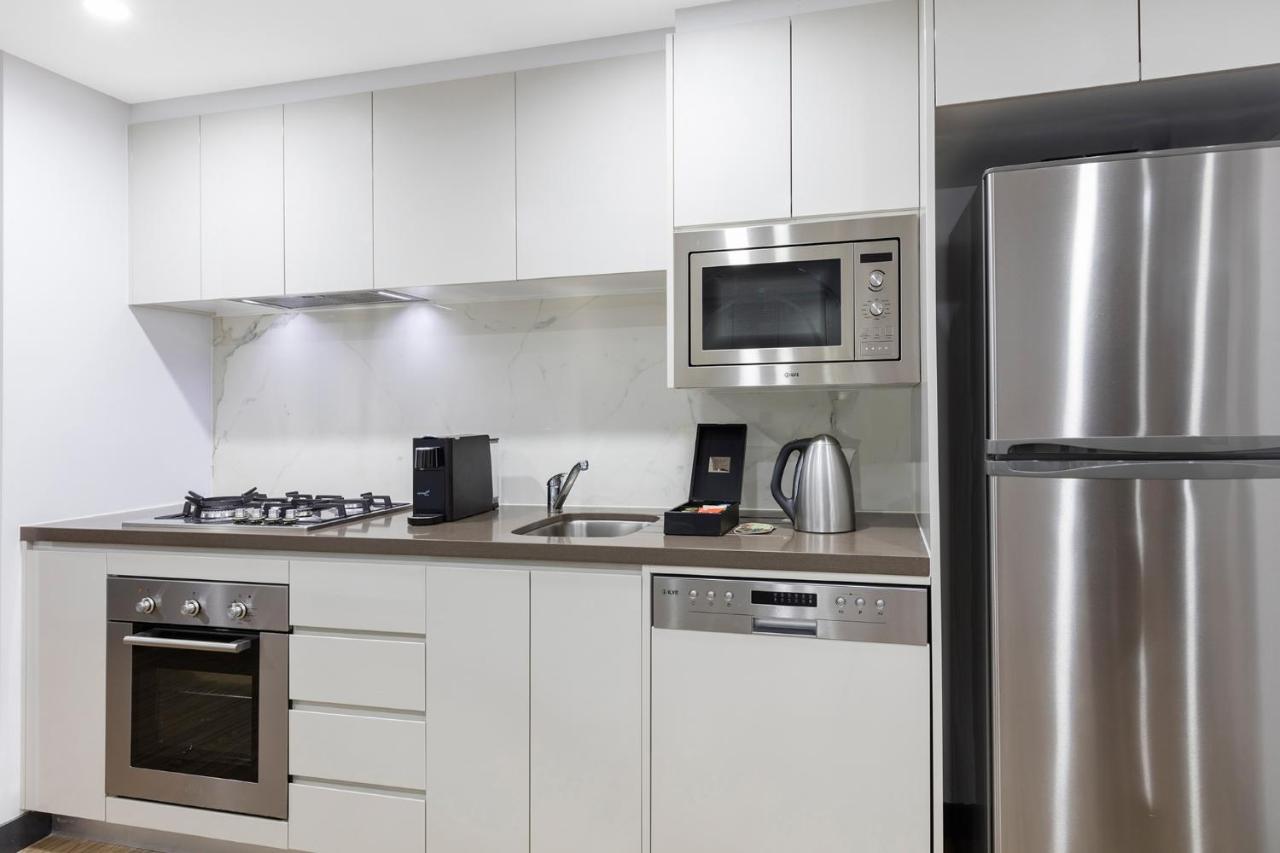 Meriton Suites Bondi Junction Сидней Экстерьер фото The photo shows a modern kitchen with a sleek, minimalist design. It features white cabinetry and a countertop that blends with the overall aesthetic. 

Key elements include:

- A stainless steel refrigerator on the right.
- An oven with a gas cookto