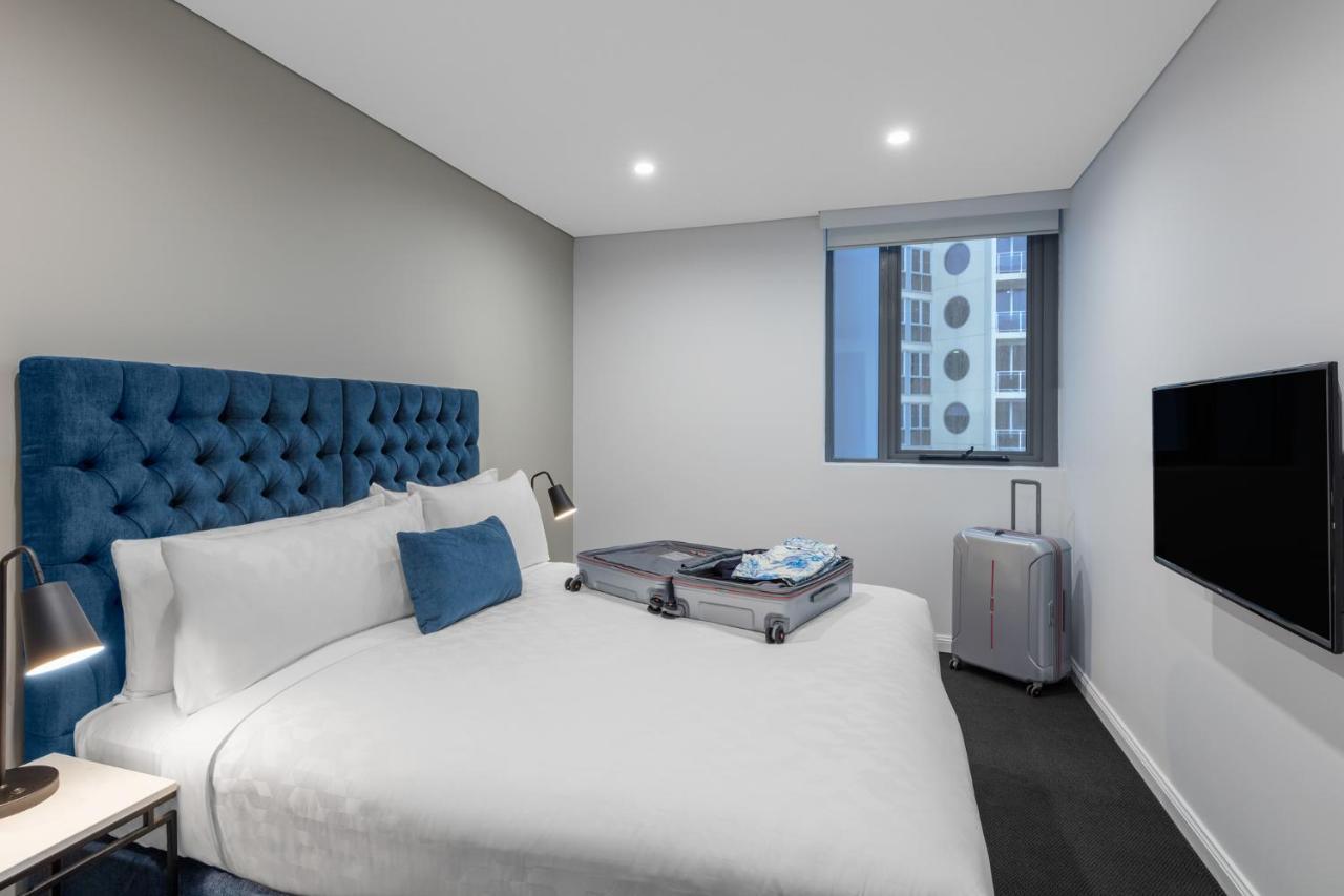Meriton Suites Bondi Junction Сидней Экстерьер фото The photo shows a modern hotel room featuring a neatly made bed with a blue tufted headboard. There is a small blue decorative pillow on the bed, and the bedding is white. Next to the bed, there is a nightstand with a lamp. A suitcase is partially op