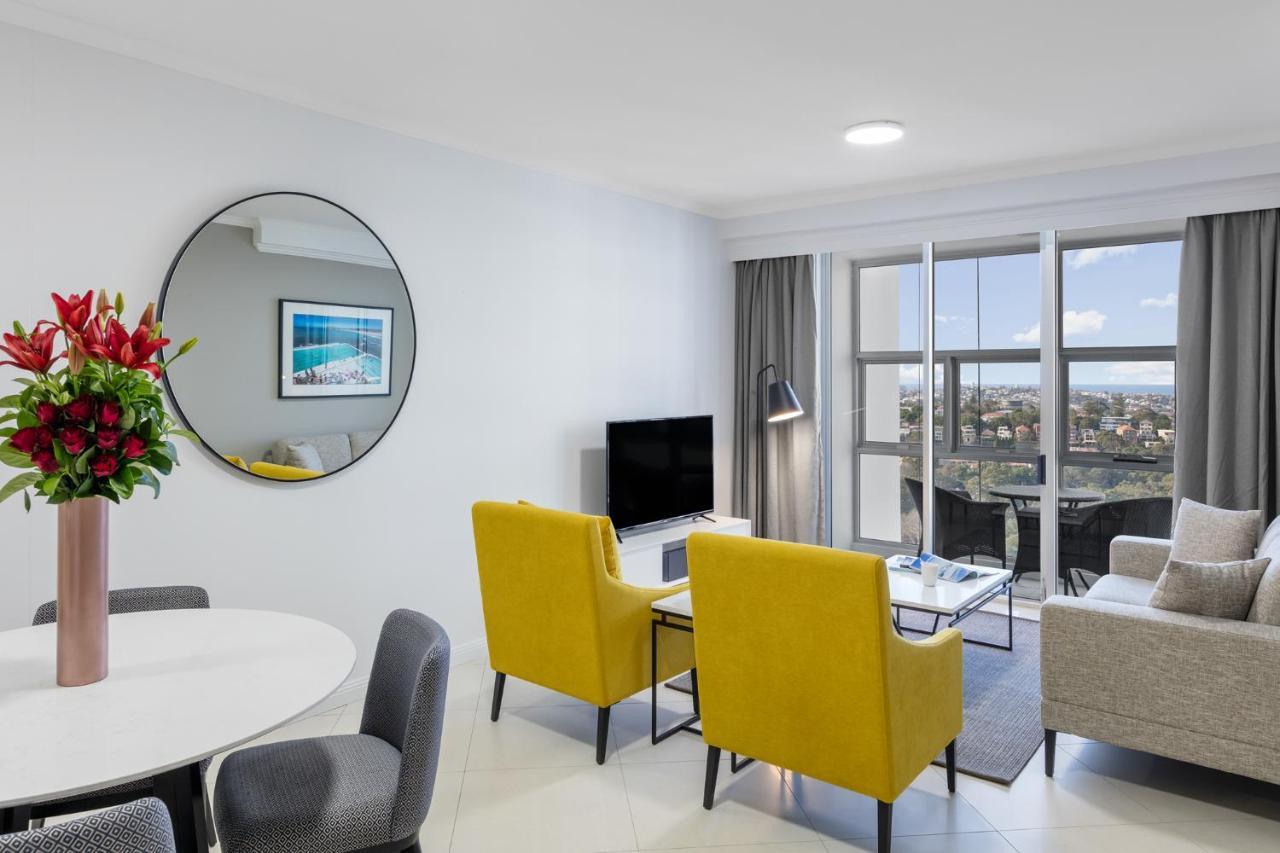 Meriton Suites Bondi Junction Сидней Экстерьер фото The photo shows a modern living space with a stylish design. There are two bright yellow chairs facing a small glass table, and a comfortable beige sofa to the right. A TV is positioned on the wall in front of the chairs. To the left, there's a dinin