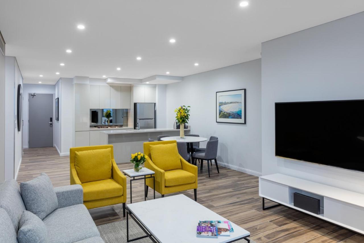 Meriton Suites Bondi Junction Сидней Экстерьер фото The photo shows a modern living space that includes a cozy sitting area and a dining area. The sitting area features two bright yellow chairs and a light gray sofa, arranged around a coffee table. There are decorative items on the table, including fl