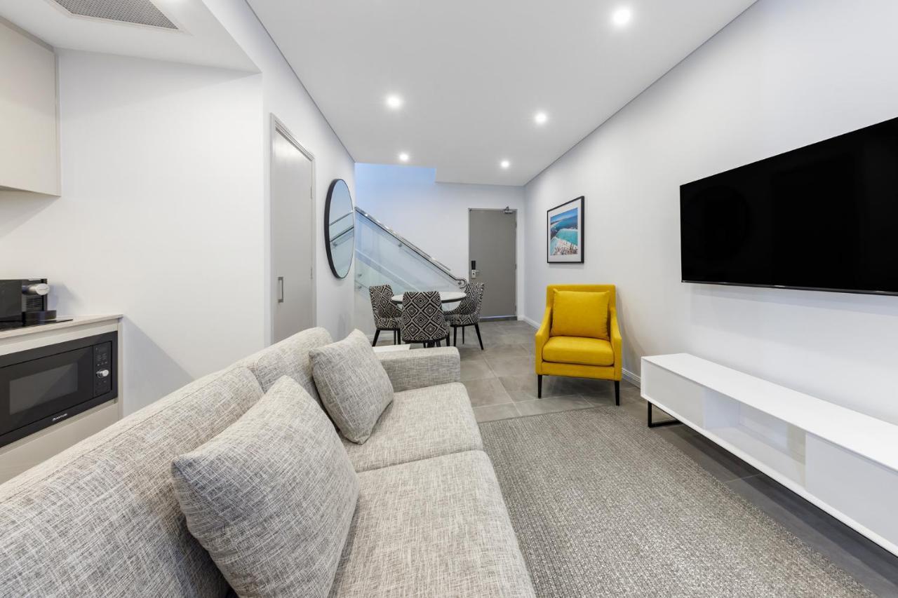Meriton Suites Bondi Junction Сидней Экстерьер фото The photo shows a modern and stylish living space. It features a light-colored sofa with cushions, positioned to face a wall-mounted flat-screen TV. There's a yellow accent chair nearby that adds a pop of color to the room. The décor is minimalistic,