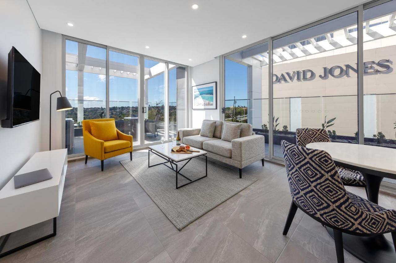 Meriton Suites Bondi Junction Сидней Экстерьер фото The photo shows a modern living space that features large windows allowing ample natural light. The room is furnished with a gray sofa and a bright yellow armchair, along with a coffee table in between. There’s a decorative bowl with fruits on the ta