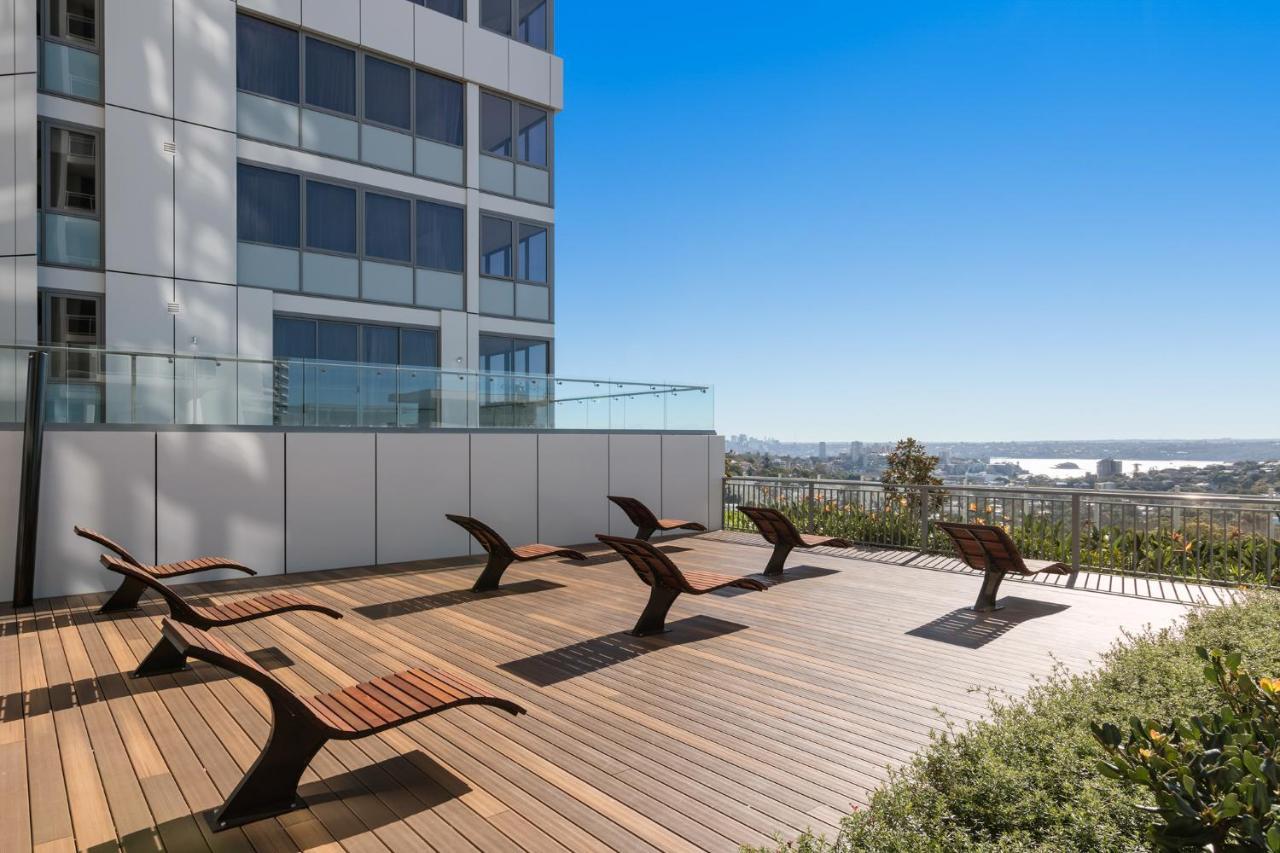Meriton Suites Bondi Junction Сидней Экстерьер фото The photo shows a rooftop or outdoor area with a wooden deck. There are several modern lounge chairs arranged neatly, facing a clear sky and a panoramic view of the surrounding landscape. In the background, there is a tall building with large windows