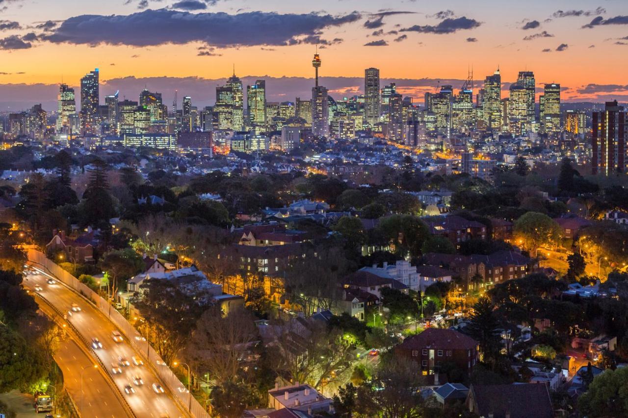 Meriton Suites Bondi Junction Сидней Экстерьер фото The photo captures a stunning city skyline at dusk, featuring towering skyscrapers adorned with lights against a colorful sunset backdrop. In the foreground, a green area with trees and houses is visible, while a road with cars can be seen winding th