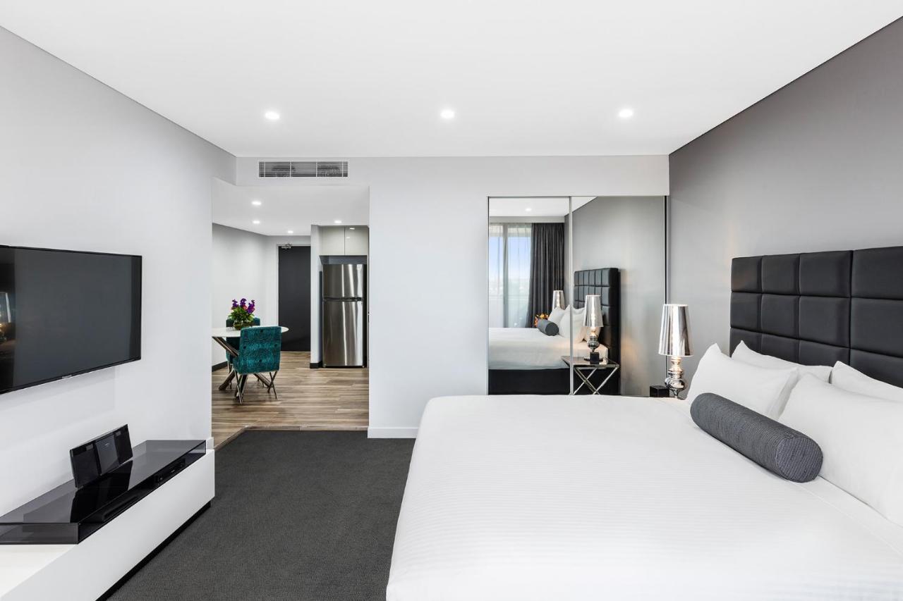 Meriton Suites Bondi Junction Сидней Экстерьер фото The photo shows a modern hotel room. It features a large bed with a white covering and gray accent pillows. To the left, there's a flat-screen TV mounted on the wall above a sleek white cabinet. The flooring is a light wood tone, and a large mirror i