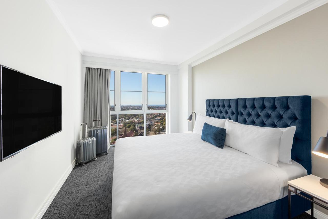 Meriton Suites Bondi Junction Сидней Экстерьер фото The photo shows a modern hotel room with a large bed featuring a tufted blue headboard. There is a white duvet and a decorative pillow on the bed. A flat-screen television is mounted on the wall opposite the bed. Large windows are covered with gray c