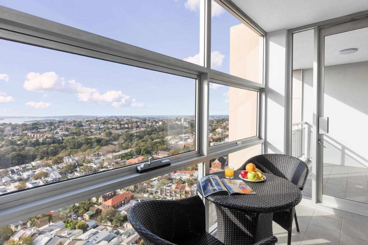 Meriton Suites Bondi Junction Сидней Экстерьер фото The photo shows a modern balcony with large glass windows that offer a panoramic view of a sprawling urban landscape. There is a small round table made of dark wicker, accompanied by two matching chairs. On the table, there is a bowl of fruit and a m