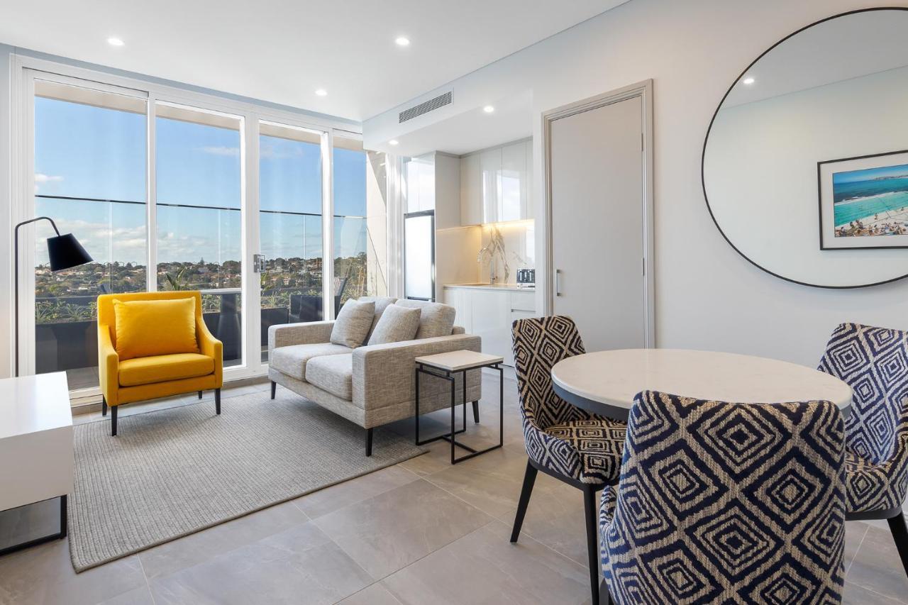 Meriton Suites Bondi Junction Сидней Экстерьер фото The photo shows a modern and stylish living area. It features a comfortable sofa in a neutral color, accompanied by a bright yellow armchair that adds a pop of color. There's a round dining table with patterned chairs nearby. Large windows allow plen