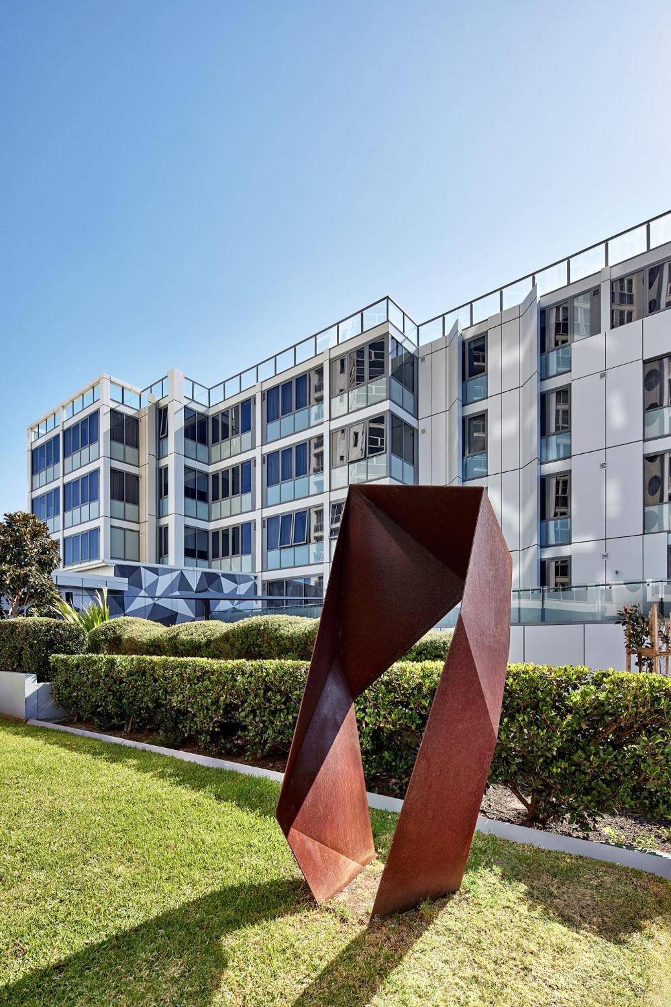 Meriton Suites Bondi Junction Сидней Экстерьер фото The photo shows a modern building with a sleek white and glass facade, featuring multiple floors and large windows. In the foreground, there is a sculptural piece made of metal, characterized by geometric shapes and a reddish-brown color. The surroun