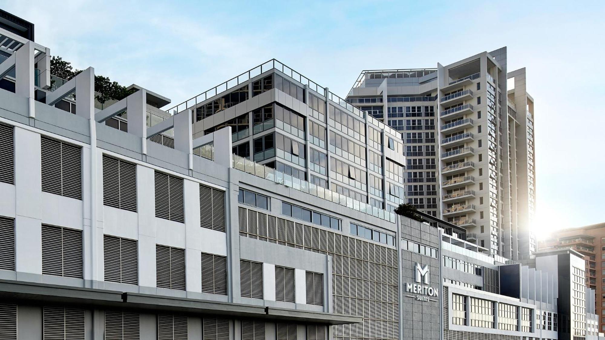 Meriton Suites Bondi Junction Сидней Экстерьер фото The image shows a modern urban building or complex. The structure features a combination of glass and concrete elements, showcasing contemporary architectural design. The foreground has a lower section that appears to be a podium or base with a textu