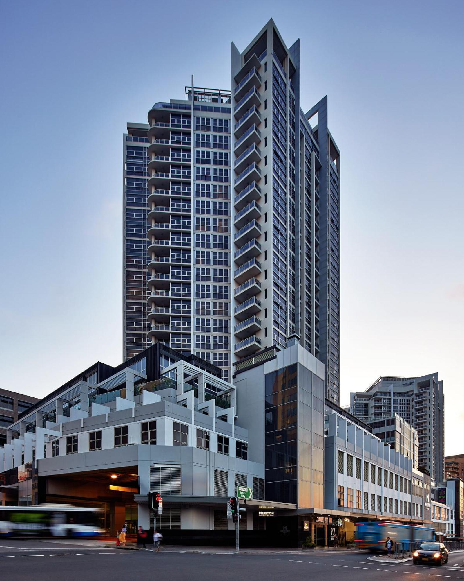 Meriton Suites Bondi Junction Сидней Экстерьер фото The image shows a modern high-rise building with a sleek design. The structure features a mix of glass and concrete elements, with multiple levels showcasing balconies. The building appears to be part of an urban environment, likely containing reside