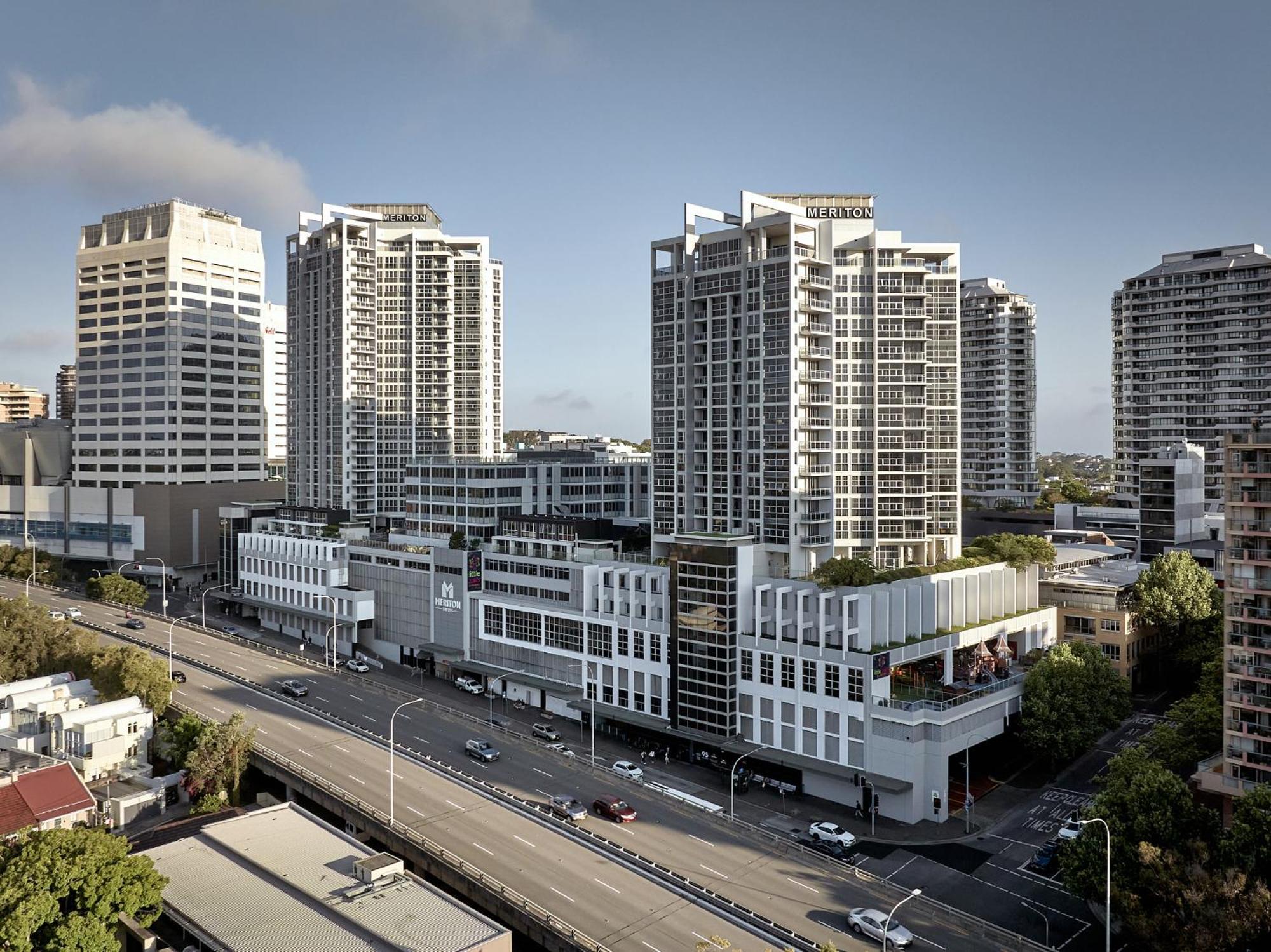 Meriton Suites Bondi Junction Сидней Экстерьер фото The photo shows a cityscape featuring several tall modern buildings, likely apartments or office complexes. In the foreground, there is a road lined with cars, and the architecture of the buildings varies, with some having sleek glass facades and oth