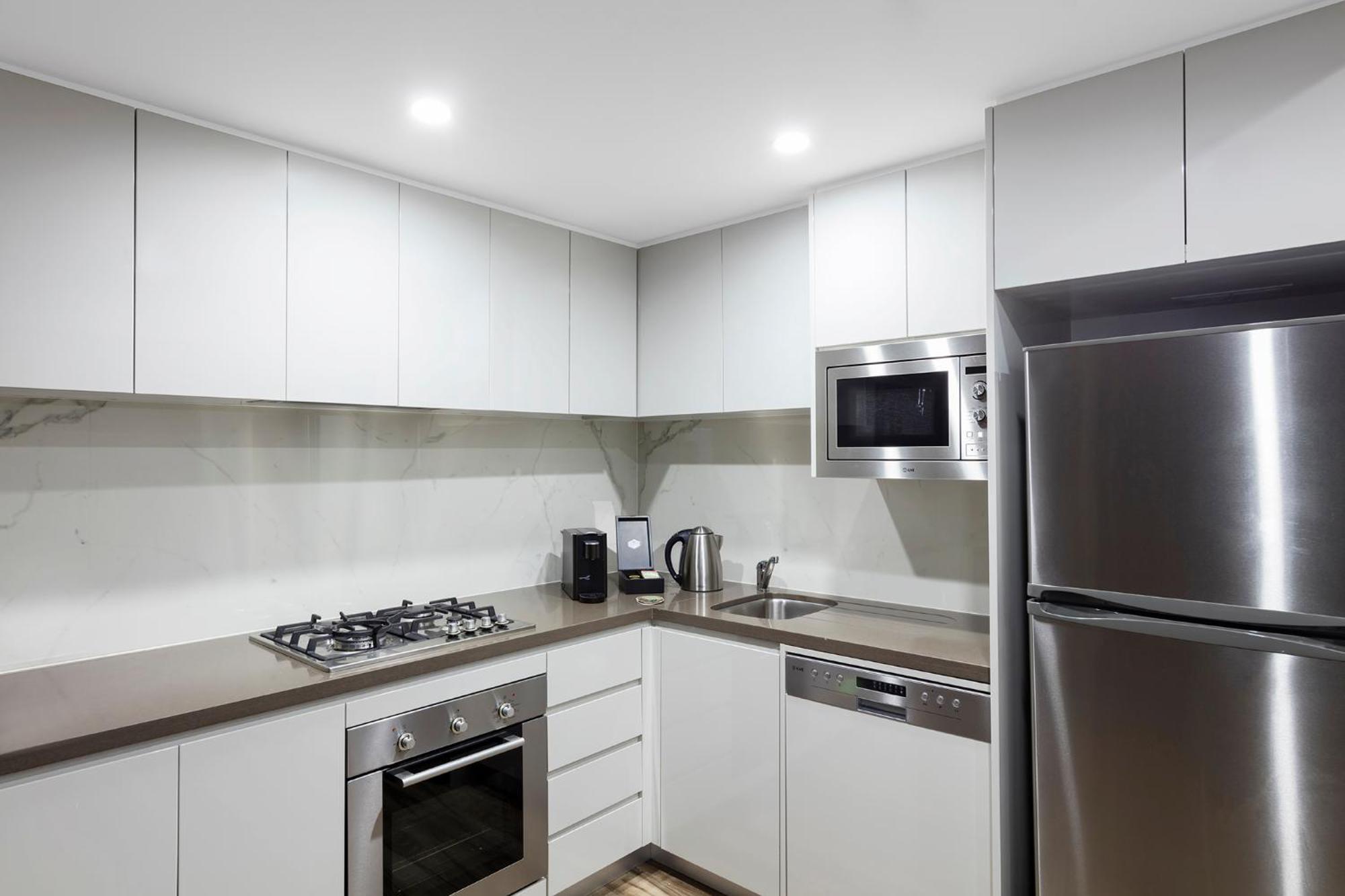 Meriton Suites Bondi Junction Сидней Экстерьер фото The photo shows a modern kitchen featuring a minimalist design. It has light-colored cabinetry, a countertop, and stainless steel appliances. The appliances include a gas stove with multiple burners, an oven, a microwave, a kettle, and a refrigerator