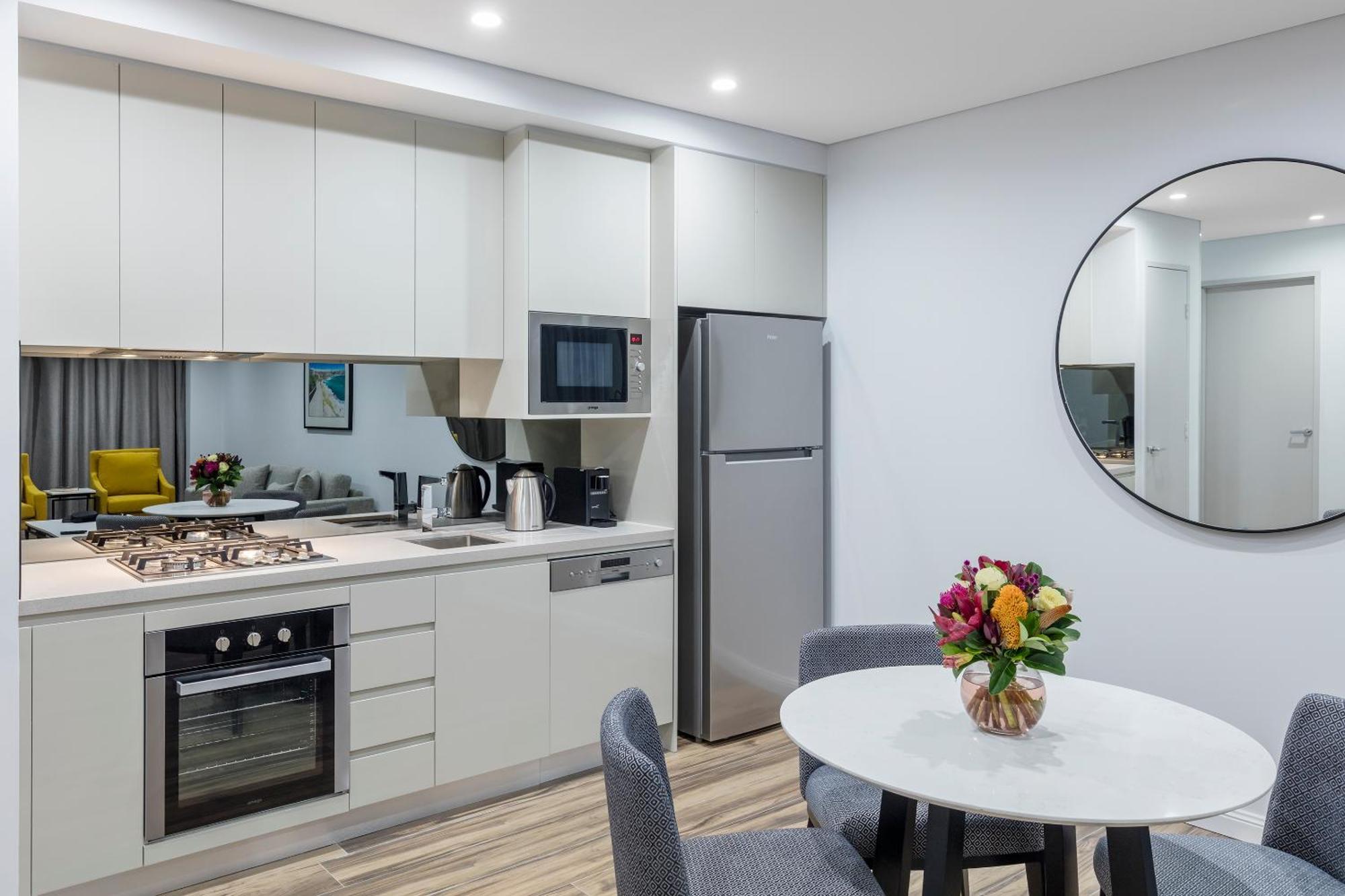 Meriton Suites Bondi Junction Сидней Экстерьер фото The photo shows a modern kitchen and dining area. The kitchen features light-colored cabinetry with a built-in oven and stovetop, a microwave, and a silver refrigerator. There is a kettle and a coffee maker on the countertop. In the dining area, ther